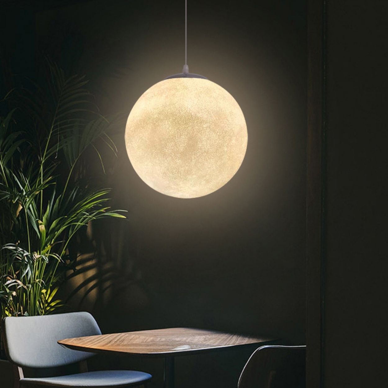 A round pendant light resembling a glowing full moon hangs above a small wooden table with two chairs. Green plants and a dark wall in the background enhance the cozy, intimate atmosphere created by the pendant light.