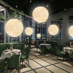 A modern restaurant interior with green chairs, round tables set for dining, large pendant lights and glowing globe lights hanging from the ceiling, and tall windows framing a nighttime city view.