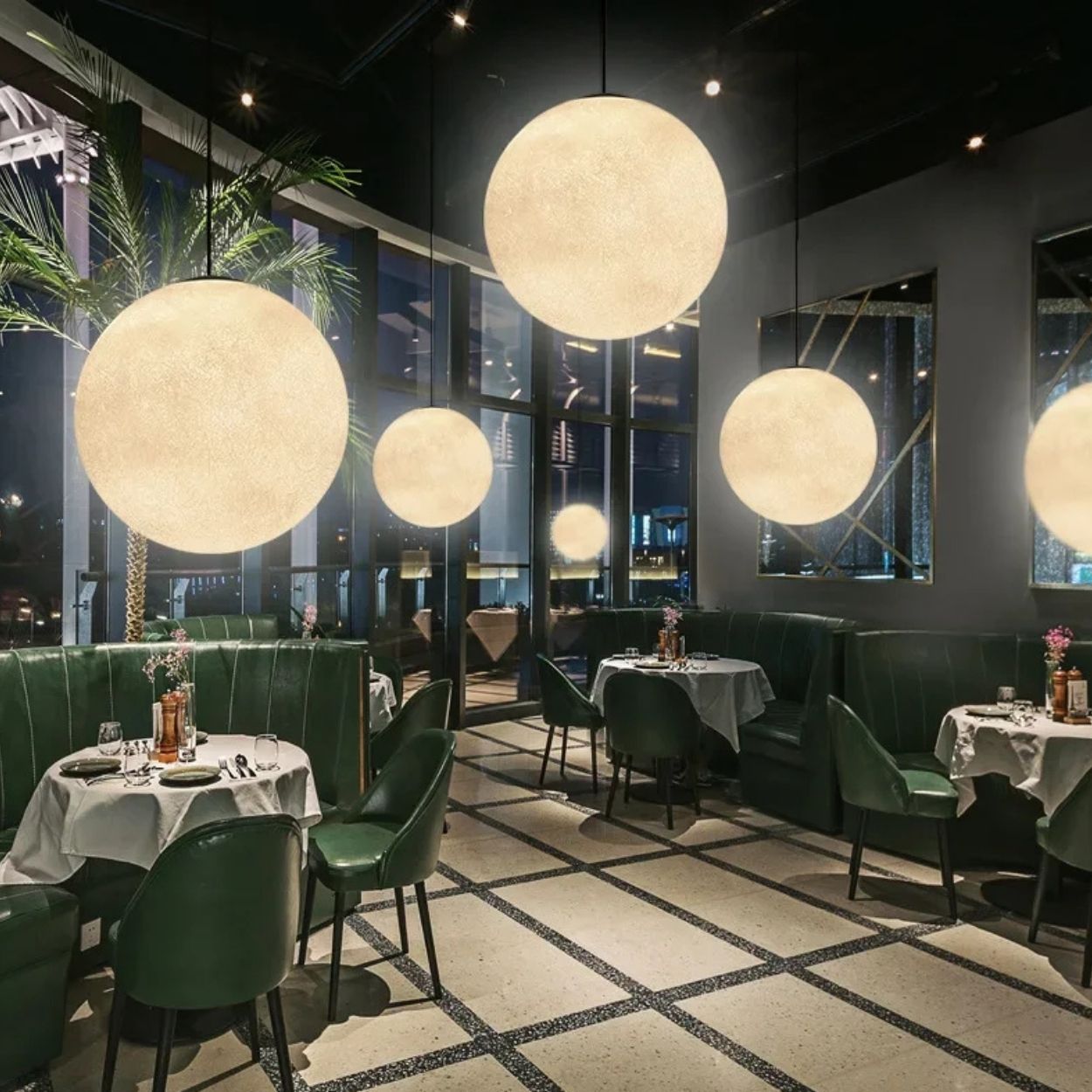 A modern restaurant interior with green chairs, round tables set for dining, large pendant lights and glowing globe lights hanging from the ceiling, and tall windows framing a nighttime city view.