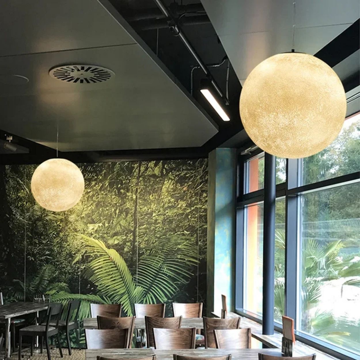 A modern dining area with large pendant lights, wooden tables and chairs, floor-to-ceiling windows, and a wall mural featuring lush green foliage.