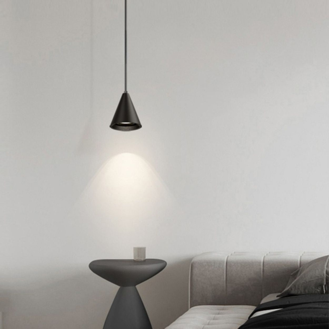 A modern, minimal bedroom with a gray upholstered bed, a small round side table, and a cone-shaped black pendant light casting light on the wall, creating a cozy and simple atmosphere.