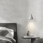 A minimal bedroom with an unmade bed, a wooden bedside table holding a silver bottle, and a cone-shaped pendant light hanging above, casting a soft glow on the light gray wall.