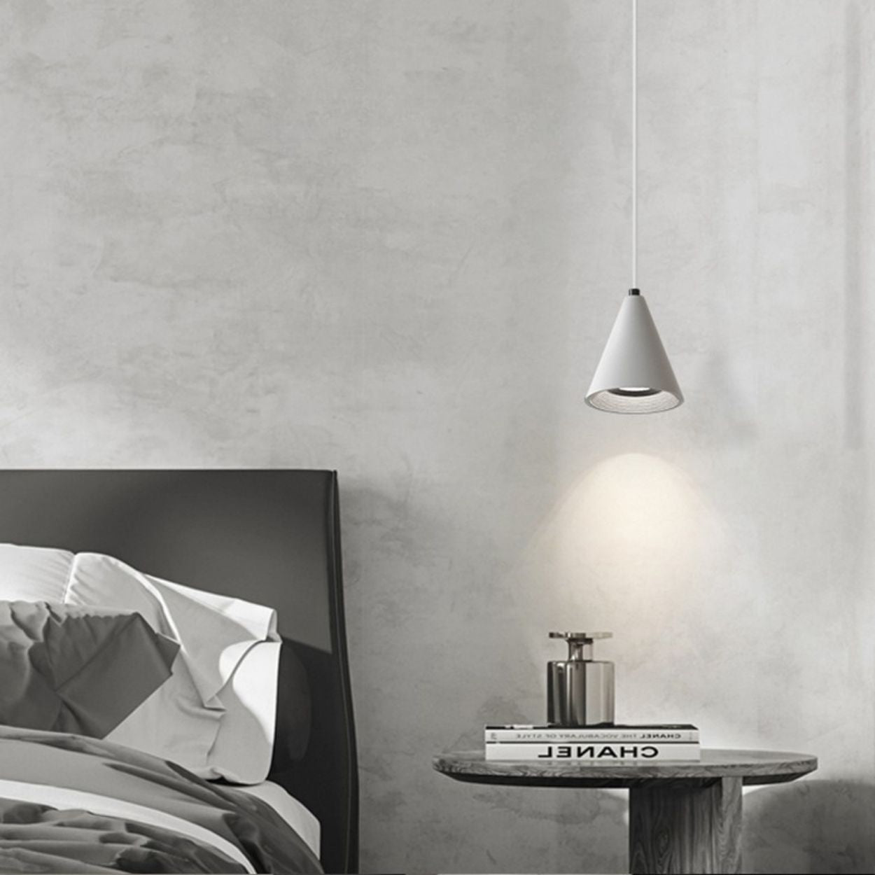 A minimal bedroom with an unmade bed, a wooden bedside table holding a silver bottle, and a cone-shaped pendant light hanging above, casting a soft glow on the light gray wall.