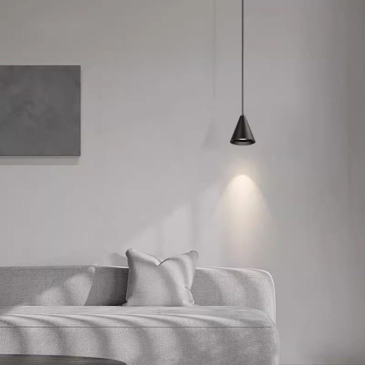 A minimal living room with a light gray sofa, a matching pillow, a blank rectangular wall art, and a single black pendant lamp casting light on the wall above. The scene is calm and uncluttered.