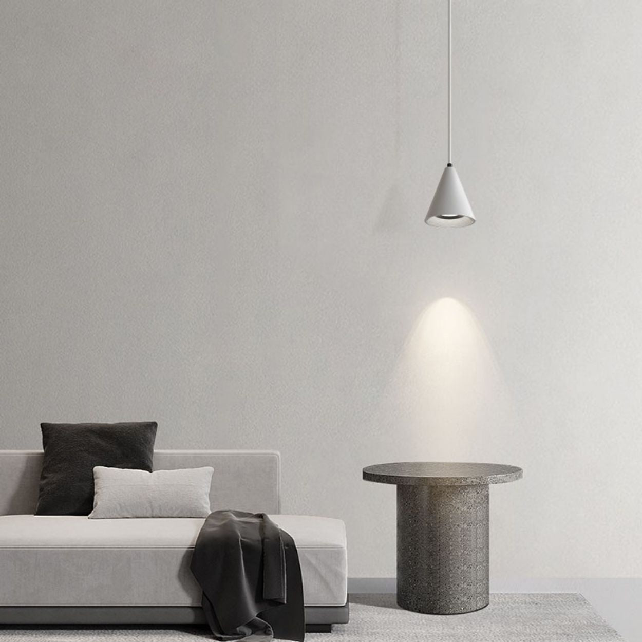 Minimalist living room with a light gray sofa, two cushions, a dark throw, a round stone side table, and a cone-shaped pendant light casting a soft glow on a neutral wall and rug.
