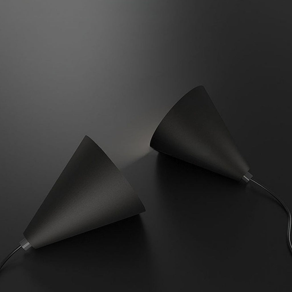 Two black, cone-shaped pendant lights lie on a dark surface, facing each other with light subtly shining from one toward the other. Both lamps have cords extending from their bases.