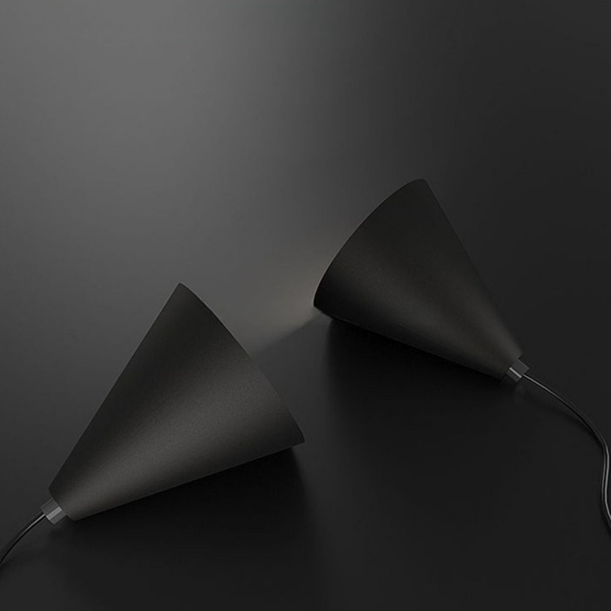 Two black, cone-shaped pendant lights lie on a dark surface, facing each other with light subtly shining from one toward the other. Both lamps have cords extending from their bases.