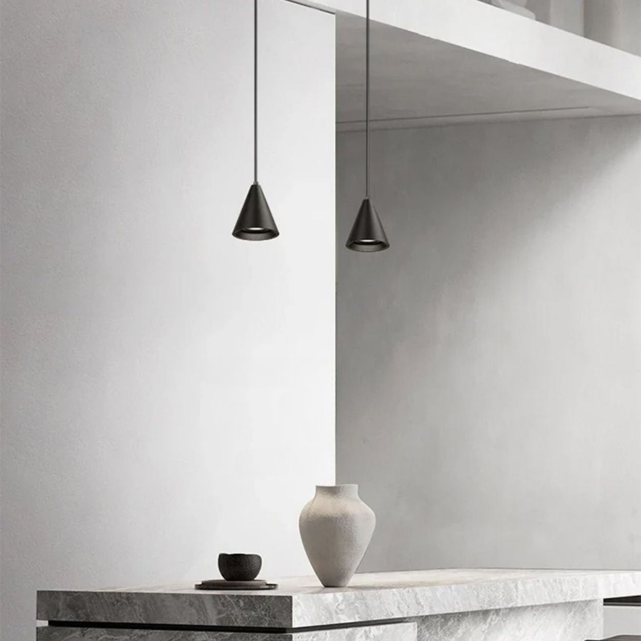 Minimalist interior with two cone-shaped black pendant lights hanging above a stone countertop, which holds a white vase and a small black cup. The background features smooth, light-colored walls.