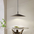 A minimalist dining area with a round table holding a plate of green olives is lit by a modern, wide-brimmed pendant lamp. Soft natural light casts gentle shadows on the light-colored wall and table.