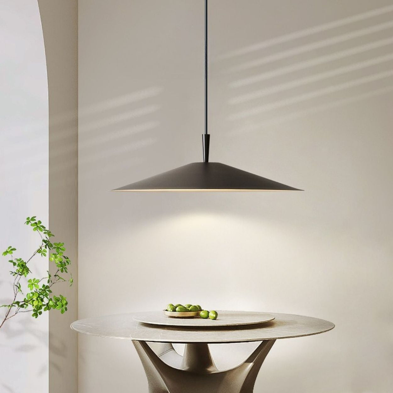 A minimalist dining area with a round table holding a plate of green olives is lit by a modern, wide-brimmed pendant lamp. Soft natural light casts gentle shadows on the light-colored wall and table.