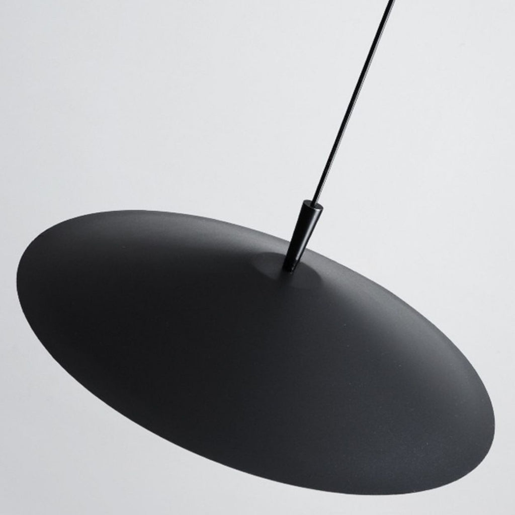 A black, round pendant light with a disc-shaped design hangs from the ceiling by a thin black cord against a plain, light gray background.