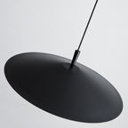 A black, round pendant light with a disc-shaped design hangs from the ceiling by a thin black cord against a plain, light gray background.
