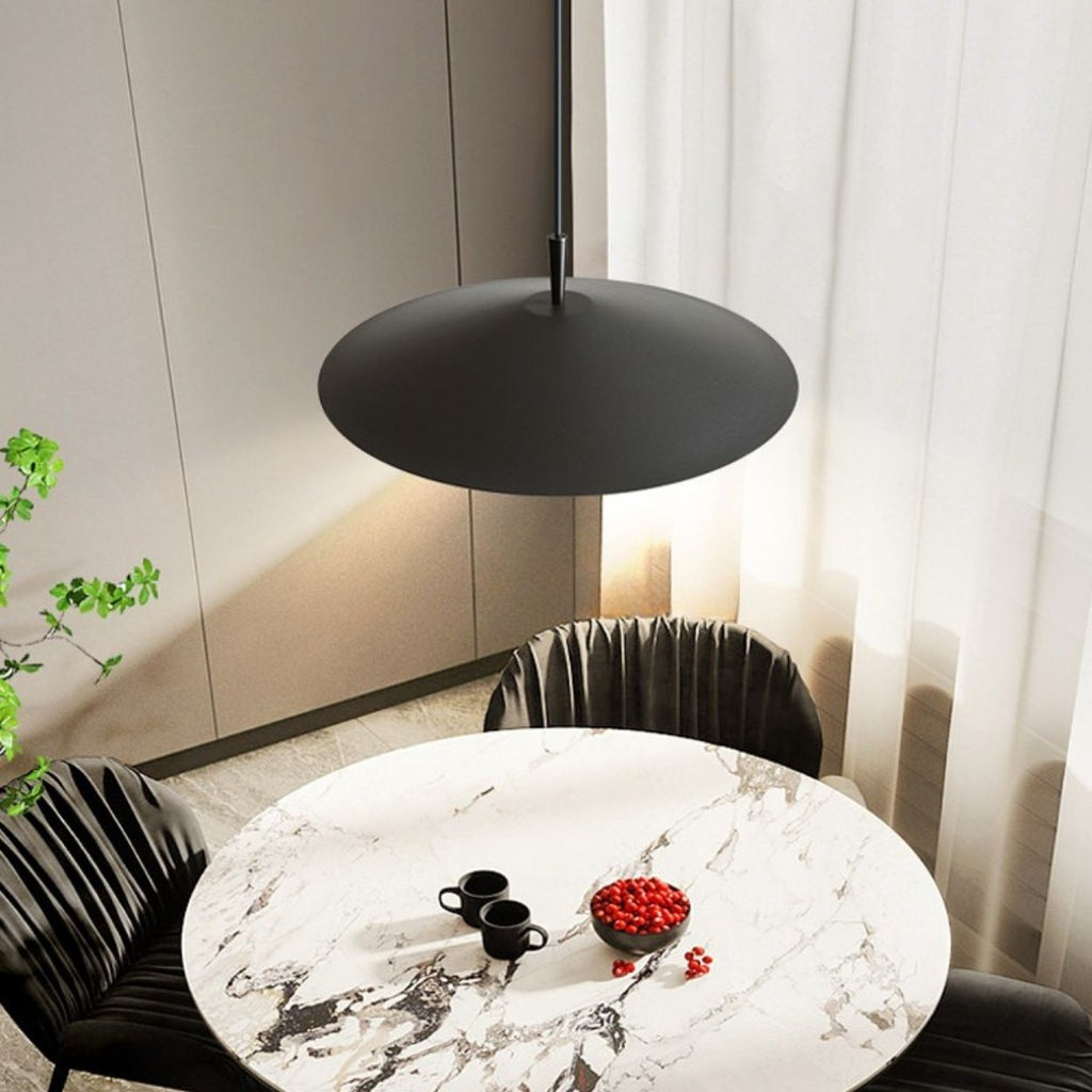A modern dining area with a round white marble table, two black chairs, a black pendant lamp above, two black cups and saucers, and a bowl of red berries, near light curtains and a green plant.