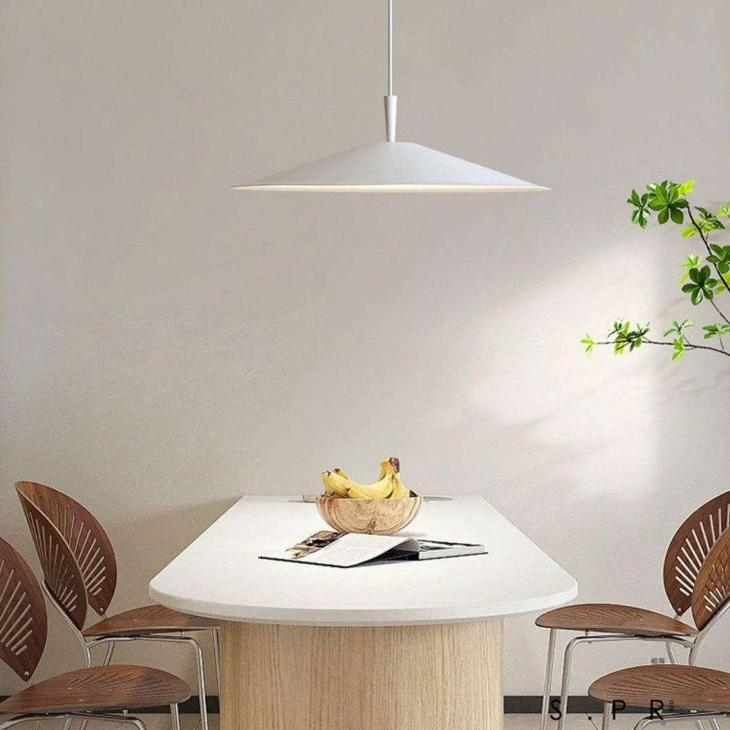 A modern dining area features a white table, four wooden chairs, a bowl of bananas, open magazines, and a striking pendant light overhead. A leafy plant sits on the right against a plain wall, adding freshness to the space.