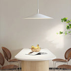 A modern dining area features a white table, four wooden chairs, a bowl of bananas, open magazines, and a striking pendant light overhead. A leafy plant sits on the right against a plain wall, adding freshness to the space.