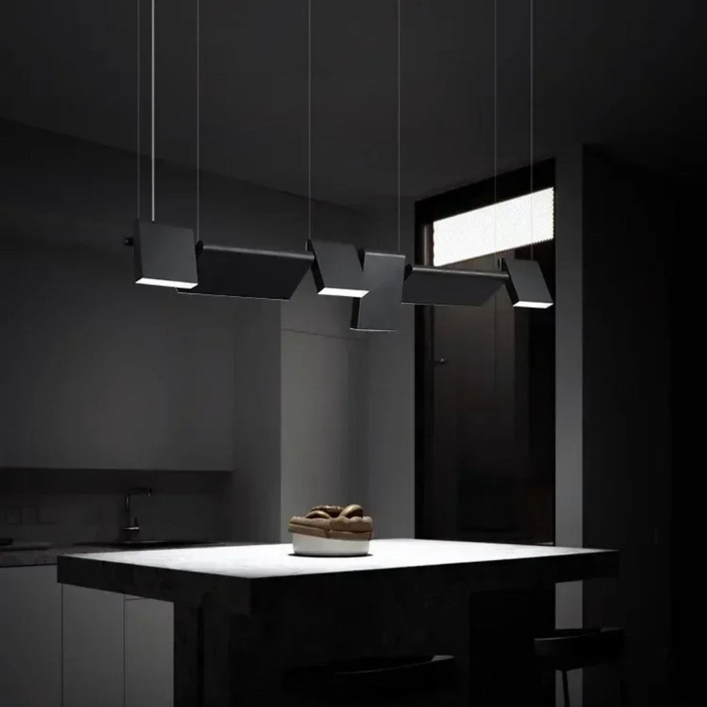 A modern kitchen with minimal lighting features a dark island countertop, a bowl of fruit, and four stylish pendant lights hanging above the island, casting focused light in an otherwise dim room.