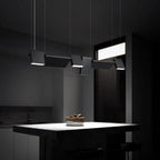 A modern kitchen with minimal lighting features a dark island countertop, a bowl of fruit, and four stylish pendant lights hanging above the island, casting focused light in an otherwise dim room.
