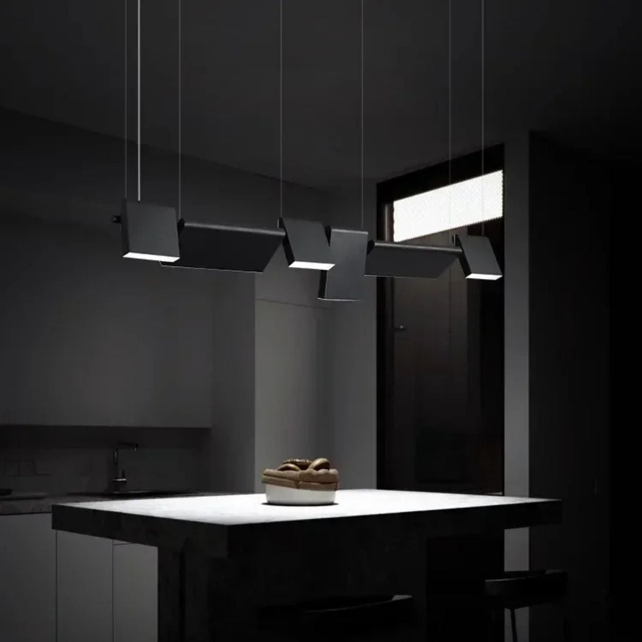 A modern kitchen with minimal lighting features a dark island countertop, a bowl of fruit, and four stylish pendant lights hanging above the island, casting focused light in an otherwise dim room.