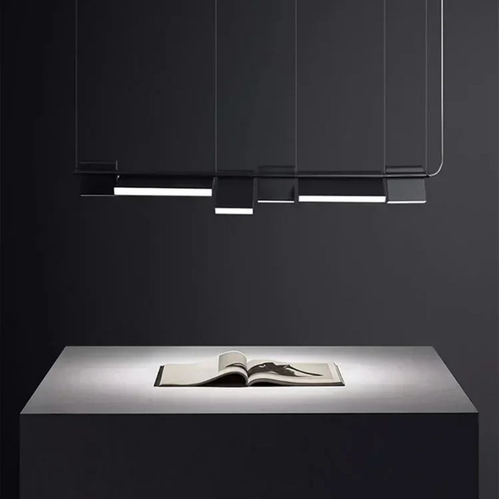A minimalist, modern pendant light with rectangular panels hangs above a sleek, gray table. An open book sits centered on the table, illuminated by the focused overhead lighting in a dark, uncluttered room.