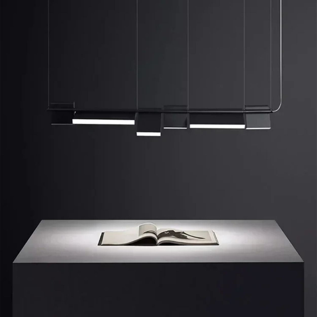 A minimalist, modern pendant light with rectangular panels hangs above a sleek, gray table. An open book sits centered on the table, illuminated by the focused overhead lighting in a dark, uncluttered room.