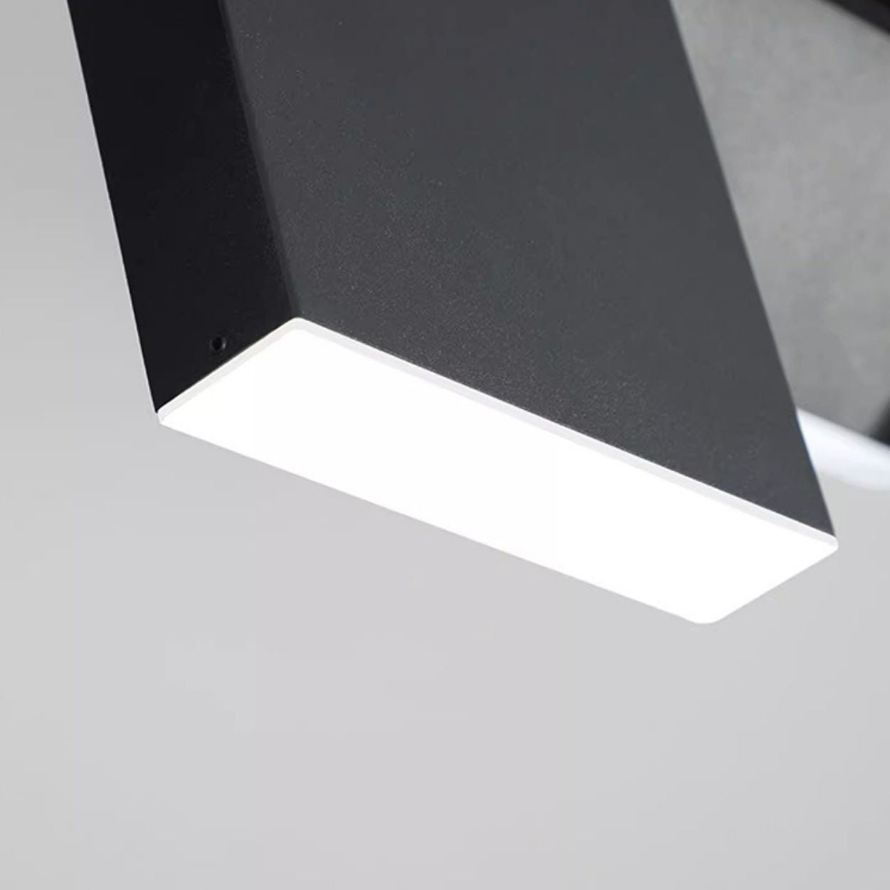 A close-up view of a modern, rectangular black pendant light fixture with a bright illuminated surface against a plain light gray background.