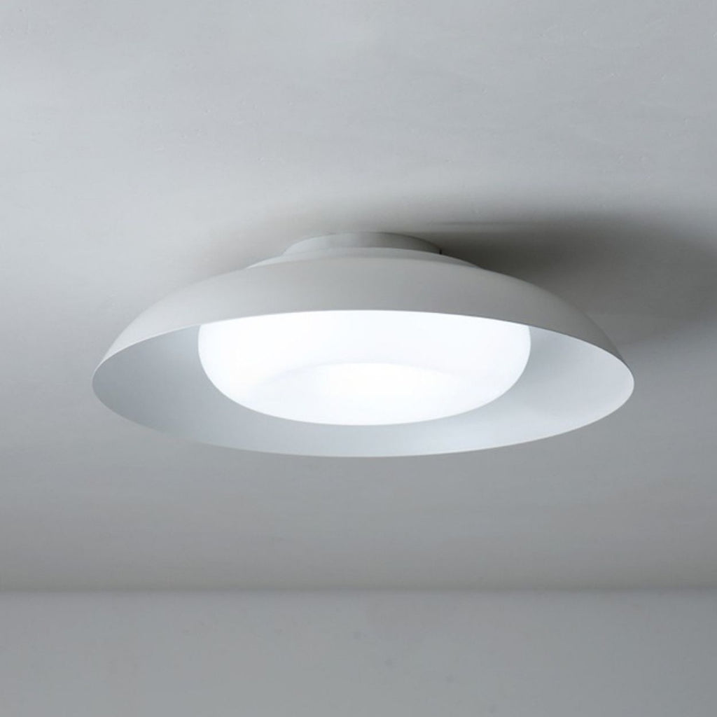 A modern, circular ceiling light fixture in white is mounted on a plain white ceiling, emitting a soft, diffused glow.