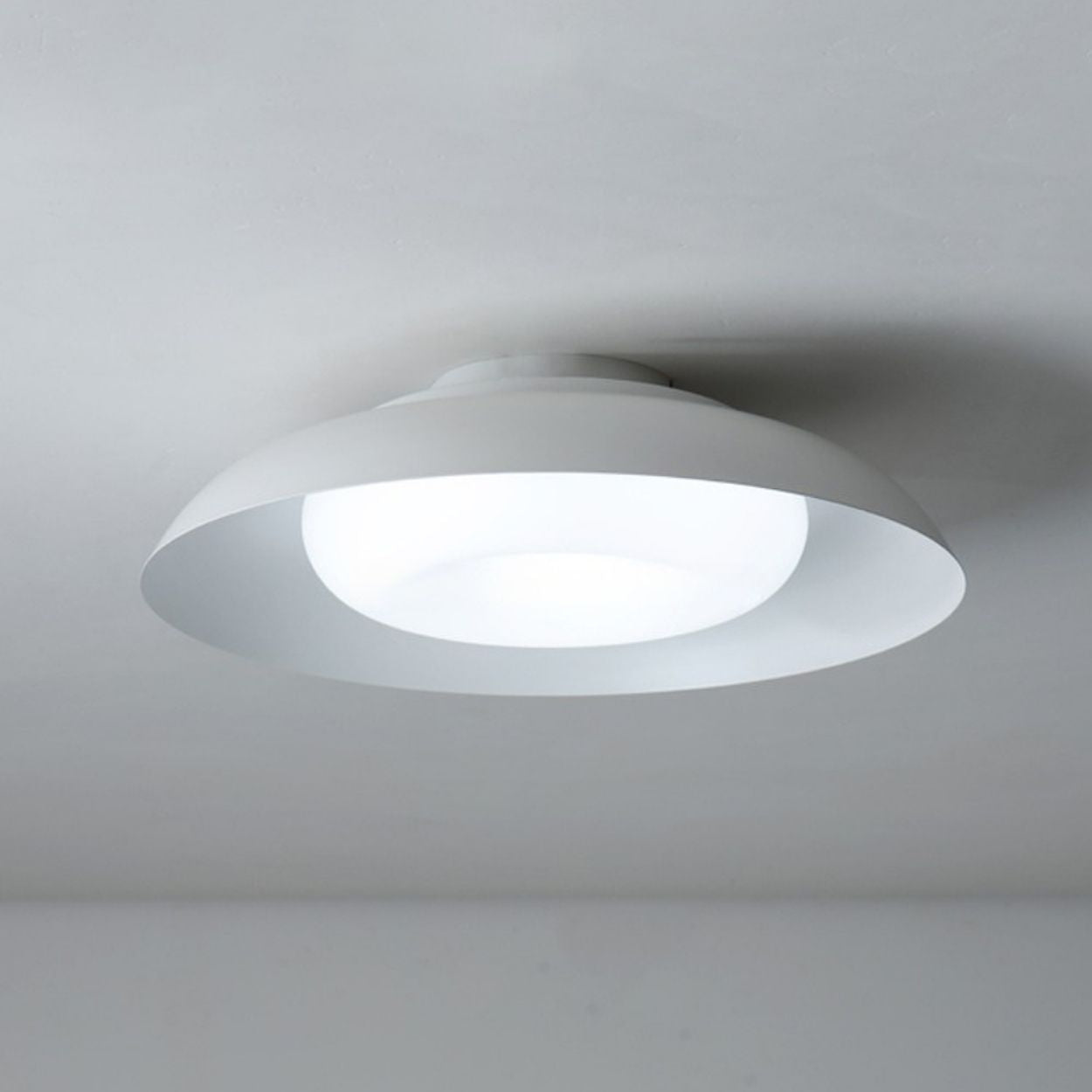 A modern, circular ceiling light fixture in white is mounted on a plain white ceiling, emitting a soft, diffused glow.