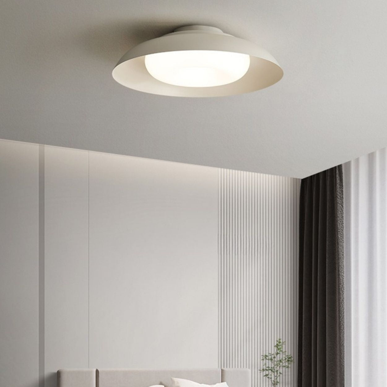 A modern ceiling light fixture emits a soft glow in a minimalist bedroom with light gray walls, a bed below, sheer curtains, and dark drapes by the window.