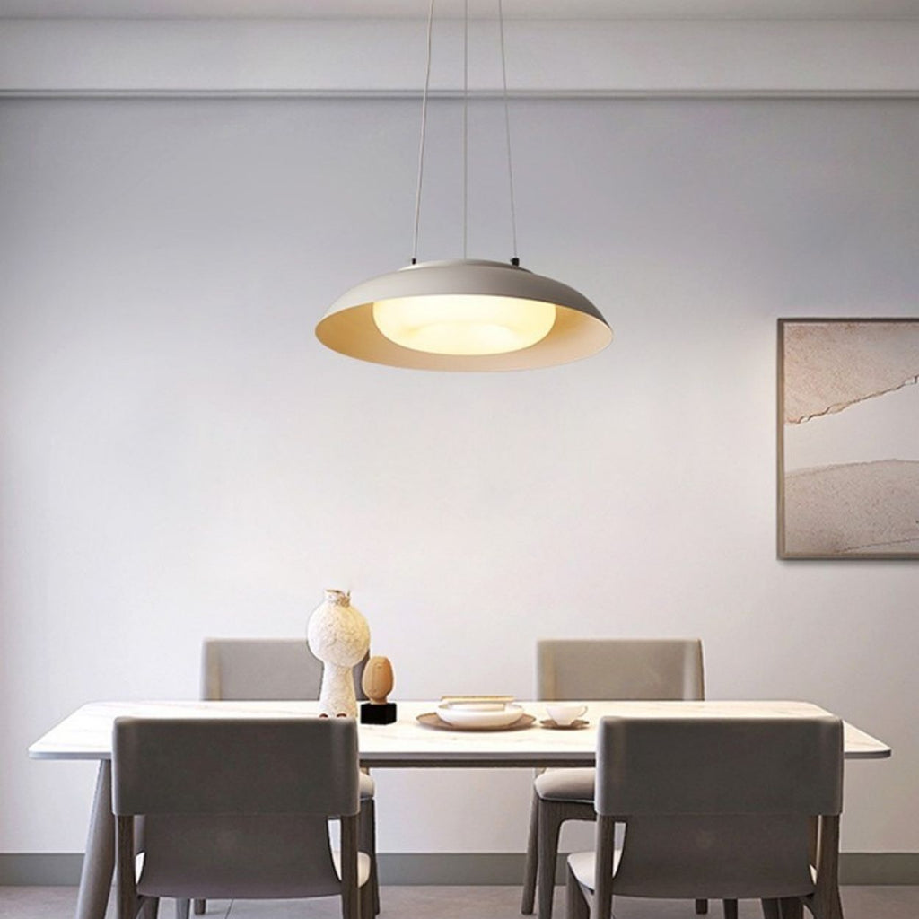 A modern dining room with a white table, four gray chairs, a stylish pendant light, minimalist decor featuring a vase, and artwork on a plain light wall.