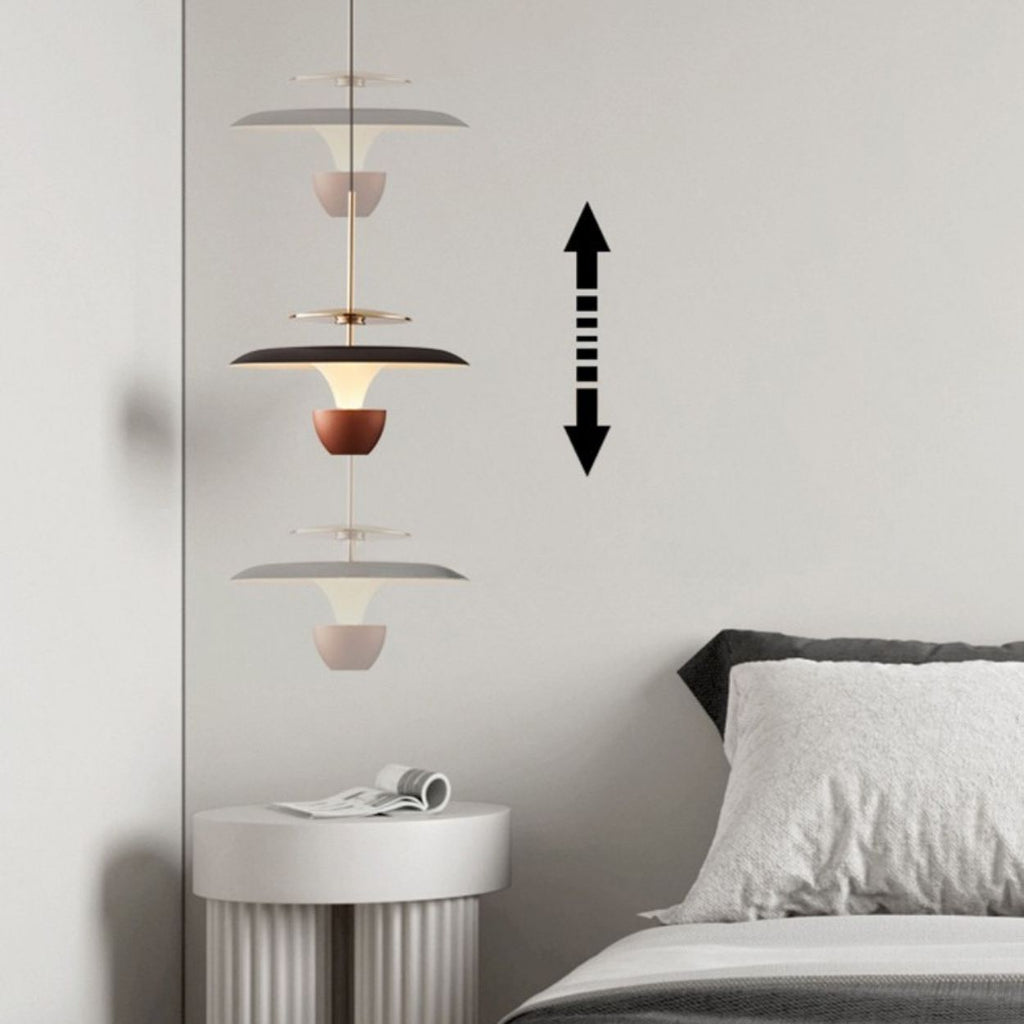 A modern luxury pendant light with layered, cone-shaped shades hangs above a white round side table next to a bed. A double-headed vertical arrow on the wall indicates the lamp’s adjustable height.