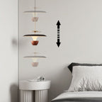 A modern luxury pendant light with layered, cone-shaped shades hangs above a white round side table next to a bed. A double-headed vertical arrow on the wall indicates the lamp’s adjustable height.