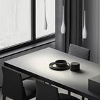 A minimalist dining room with a gray table set with black dishes, surrounded by dark chairs, large windows with sheer curtains, and sleek pendant light fixtures hanging above for a modern touch.