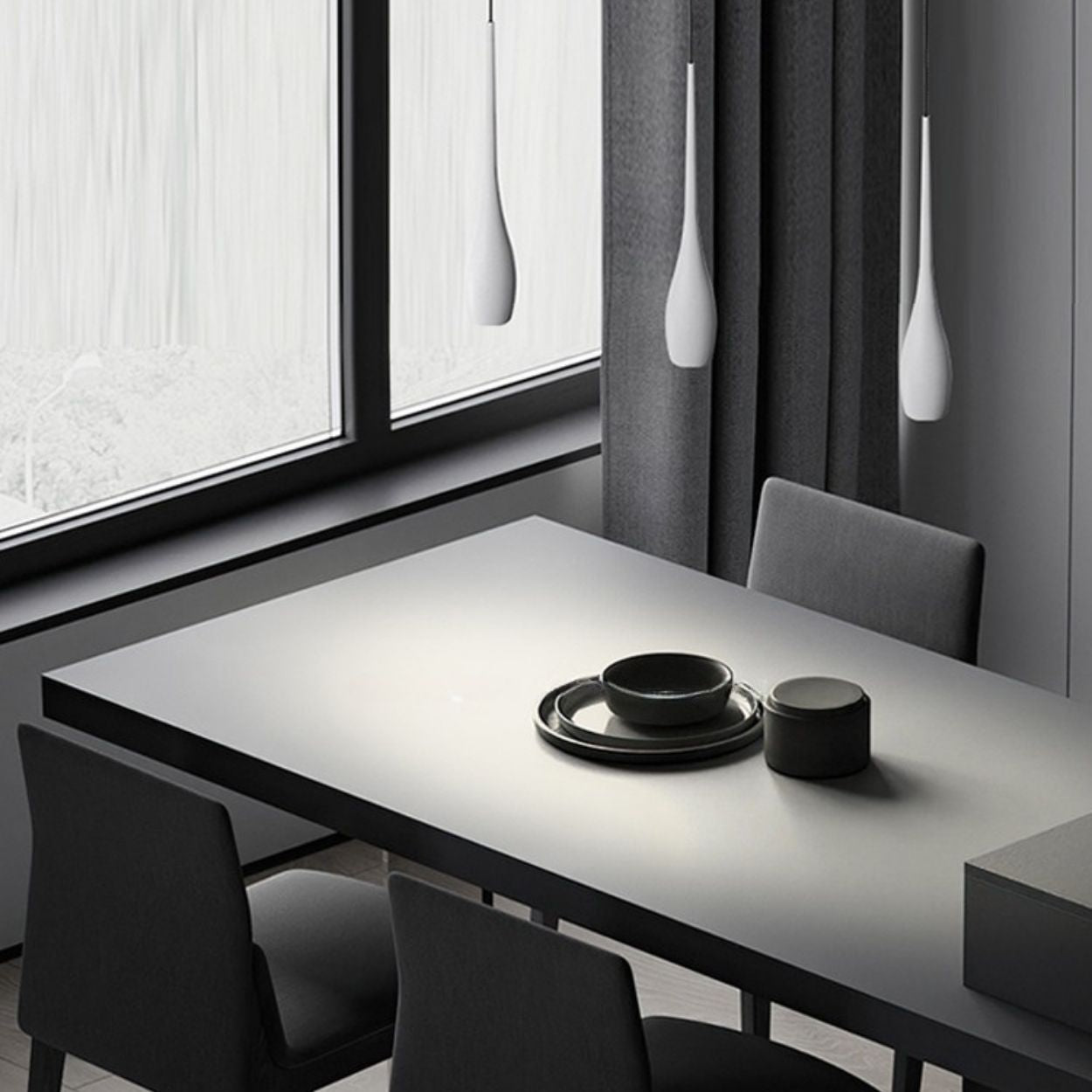 A minimalist dining room with a gray table set with black dishes, surrounded by dark chairs, large windows with sheer curtains, and sleek pendant light fixtures hanging above for a modern touch.