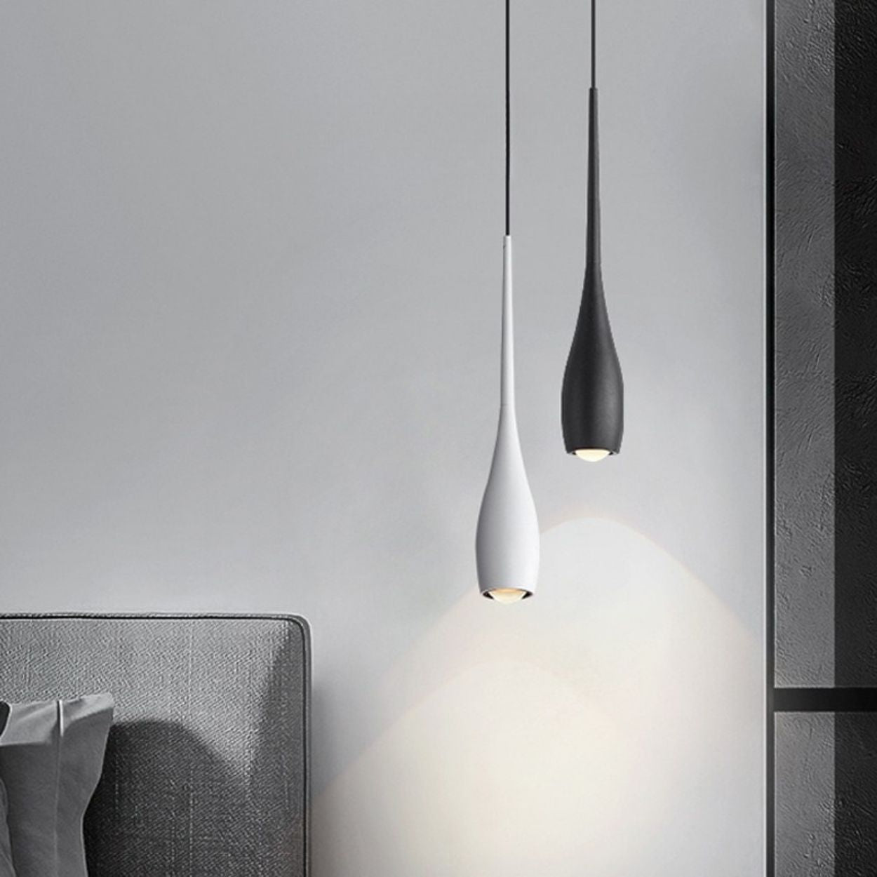Two modern pendant lights, one white and one black, hang above a gray upholstered bed and cast soft light on a light gray wall in this minimalist bedroom setting.