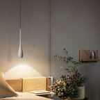 A modern, minimalistic bedroom with a hanging pendant light over a wooden nightstand, a potted plant, and a small wall shelf holding a book. The room has neutral-colored walls and bedding.