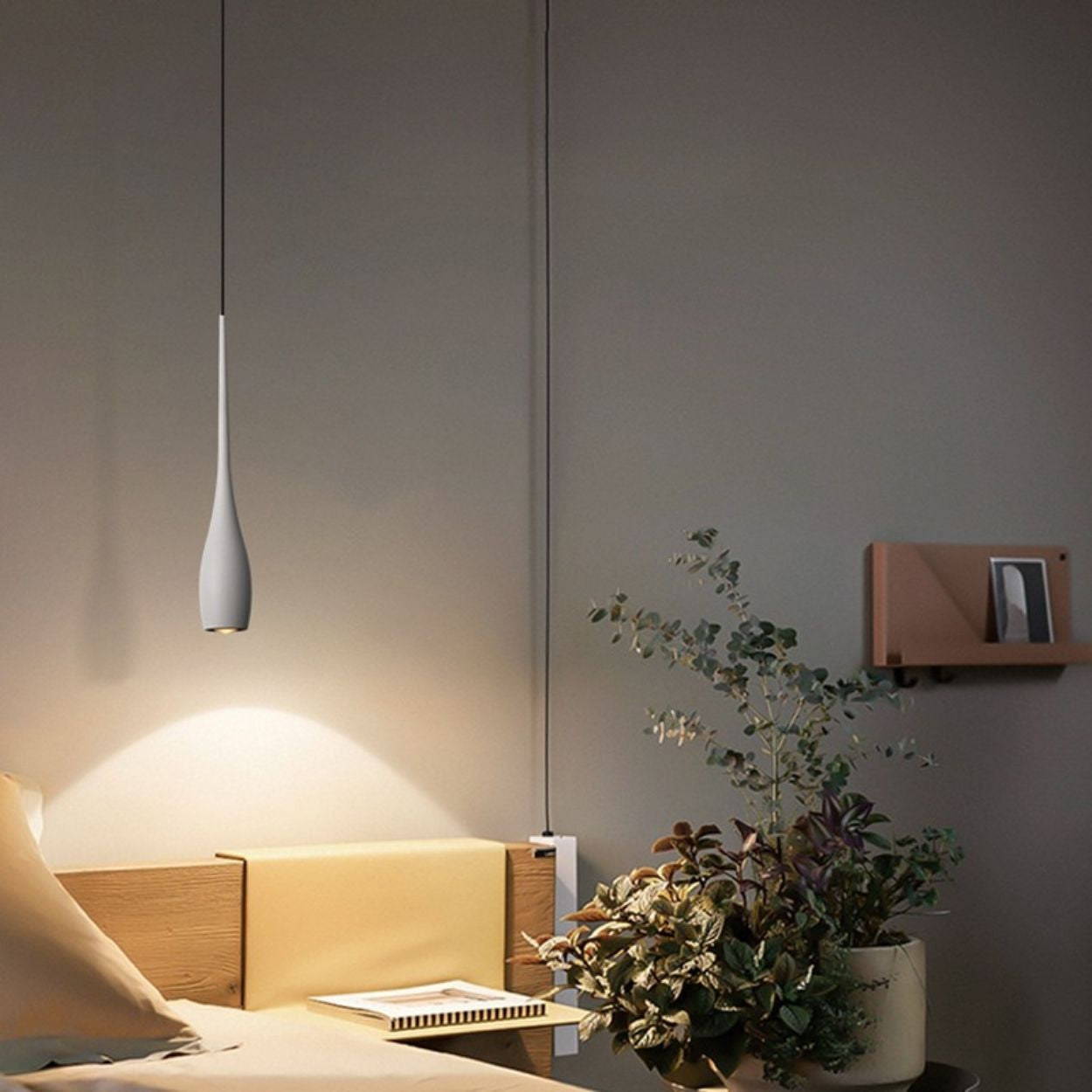 A modern, minimalistic bedroom with a hanging pendant light over a wooden nightstand, a potted plant, and a small wall shelf holding a book. The room has neutral-colored walls and bedding.