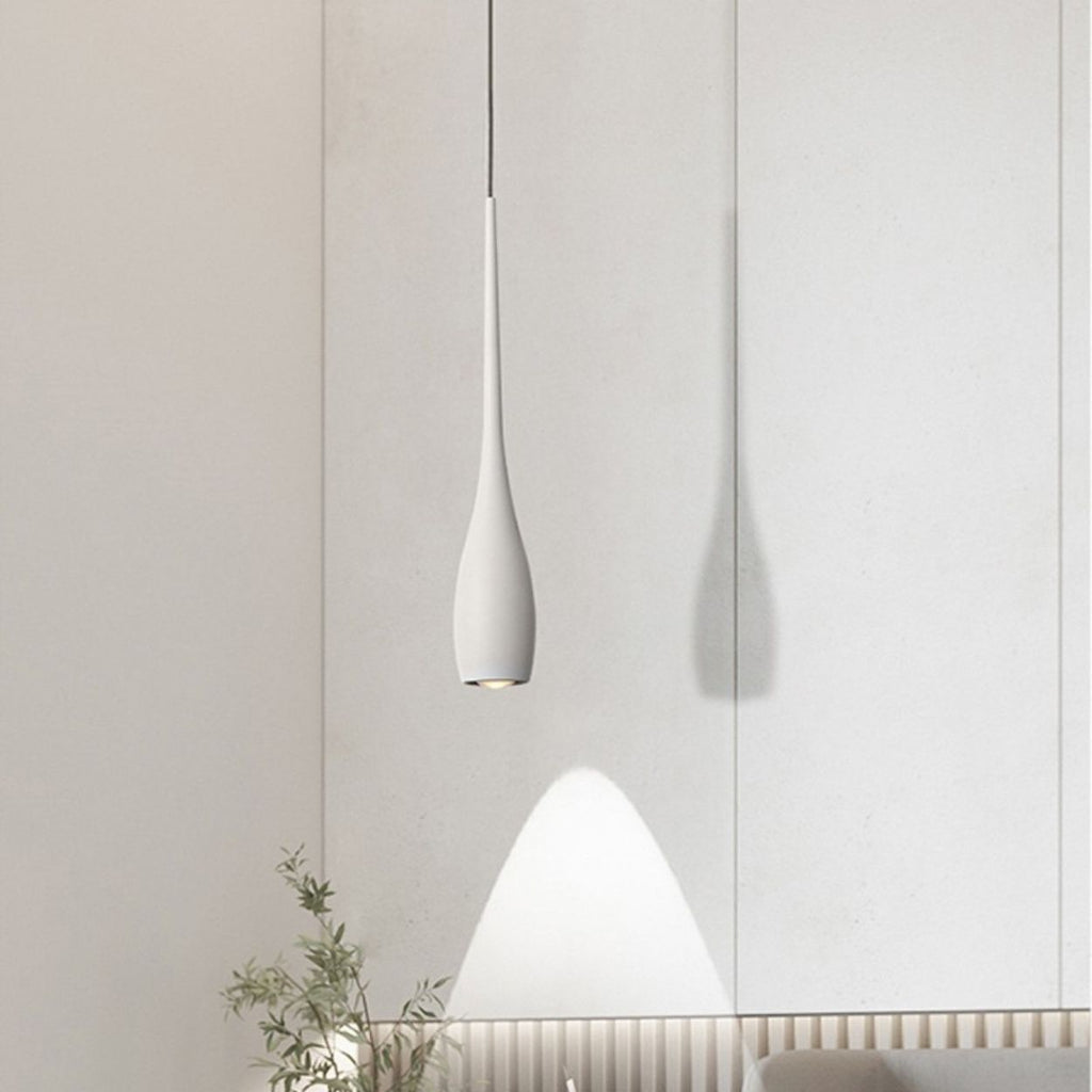 A modern, minimalist pendant light in white hangs before a light-colored wall, casting a soft shadow and a gentle cone of light. A small plant with green leaves sits in the bottom left corner, adding freshness to the scene.