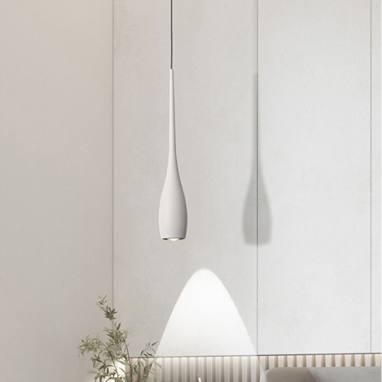 A modern, minimalist pendant light in white hangs before a light-colored wall, casting a soft shadow and a gentle cone of light. A small plant with green leaves sits in the bottom left corner, adding freshness to the scene.