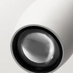 Close-up of a modern, minimalistic white pendant light with a round, reflective glass lens or surface, featuring a smooth texture and clean lines against a light gray background.