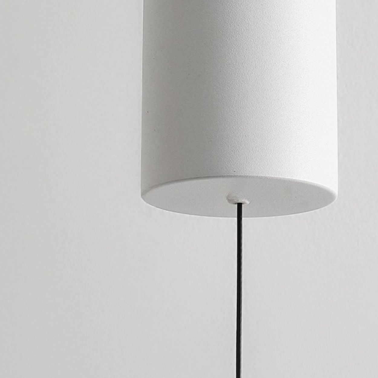 Close-up of a modern, cylindrical white pendant light mount hanging from a thin black cord against a plain light gray wall.
