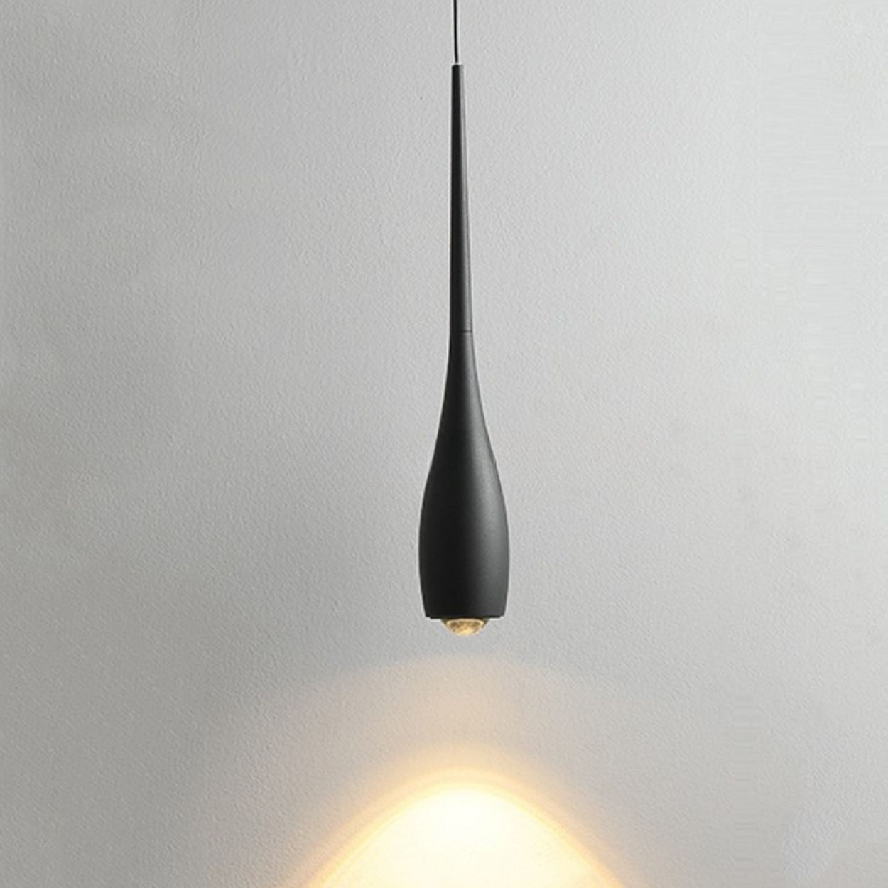 A modern black pendant light with a sleek, elongated teardrop shape hangs from the ceiling, casting a warm, focused beam of light onto a plain white wall—perfect for showcasing the elegance of contemporary pendant lighting.