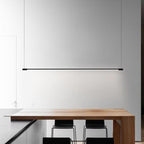 A minimal modern kitchen features a sleek wooden table with two black chairs, a black countertop, and a stylish pendant light suspended in front of a large blank white wall.