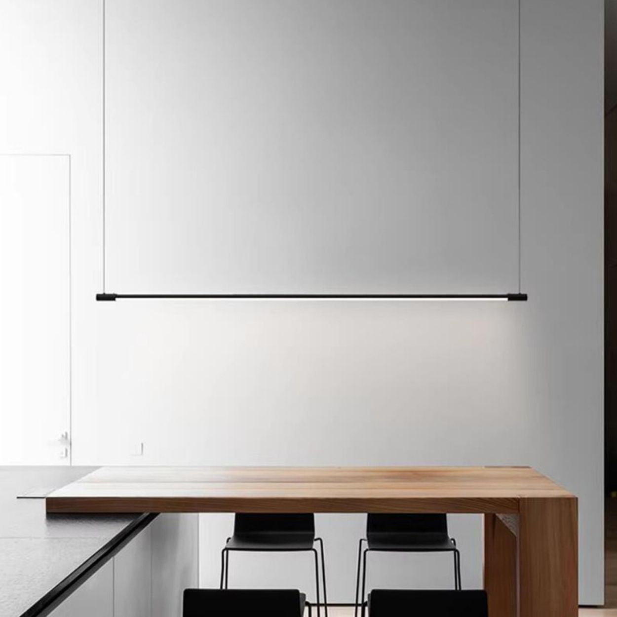 A minimal modern kitchen features a sleek wooden table with two black chairs, a black countertop, and a stylish pendant light suspended in front of a large blank white wall.