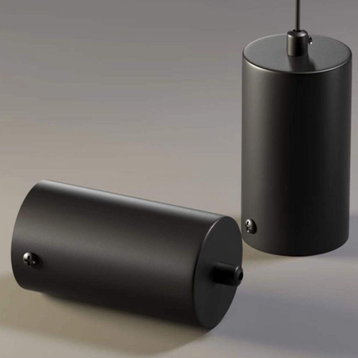 Two matte black cylindrical objects are shown in a pendant light mount close up; one stands upright with a cord attached to its top, while the other lies horizontally. Both feature small protrusions and rounded ends against a softly lit, neutral background.