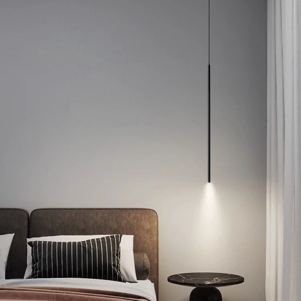 A modern bedroom with a brown upholstered bed, black and white striped pillows, a small round side table, and a sleek pendant light casting a soft glow on the table. Light gray walls and white curtains complete the serene ambiance.