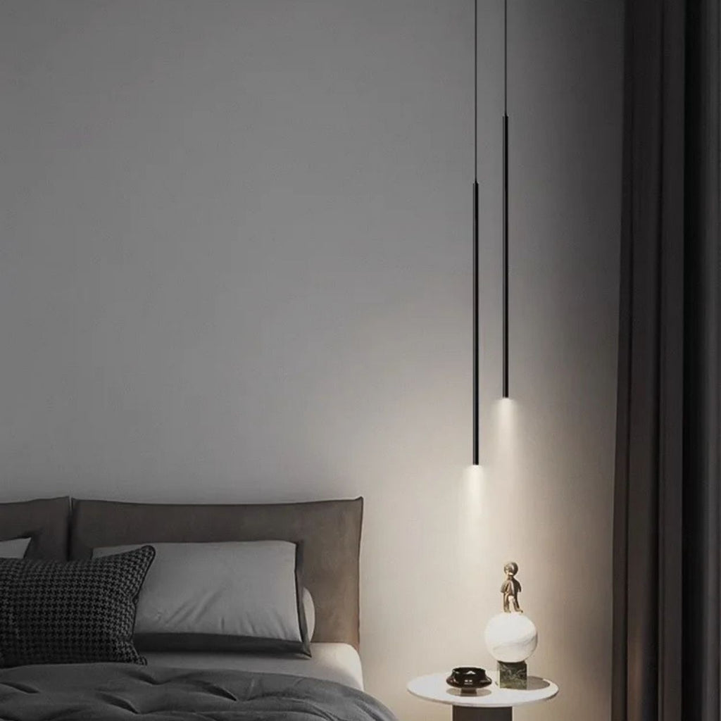 A modern bedroom features a grey bed with dark pillows, a round white side table holding a small sculpture and lamp, and two pendant lights hanging from the ceiling beside rich, dark curtains.