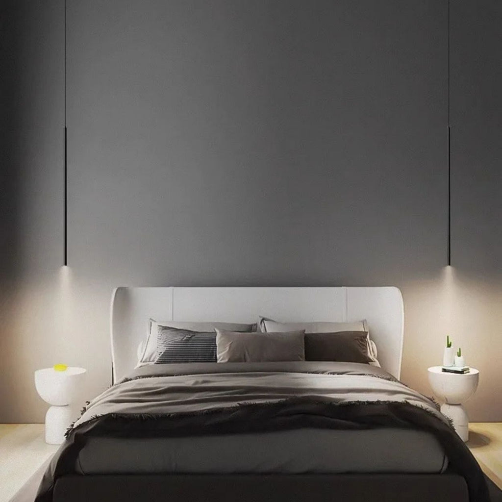 A modern bedroom features a large bed with gray and beige bedding, flanked by two white nightstands. A pendant light hangs above each nightstand, adding sleek illumination against the solid dark gray wall behind the bed.