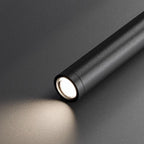 A sleek black cylindrical flashlight, resembling a cylindrical pendant light source, emits a soft beam of white light onto a smooth, dark surface, creating a gentle illuminated area in front of the device.