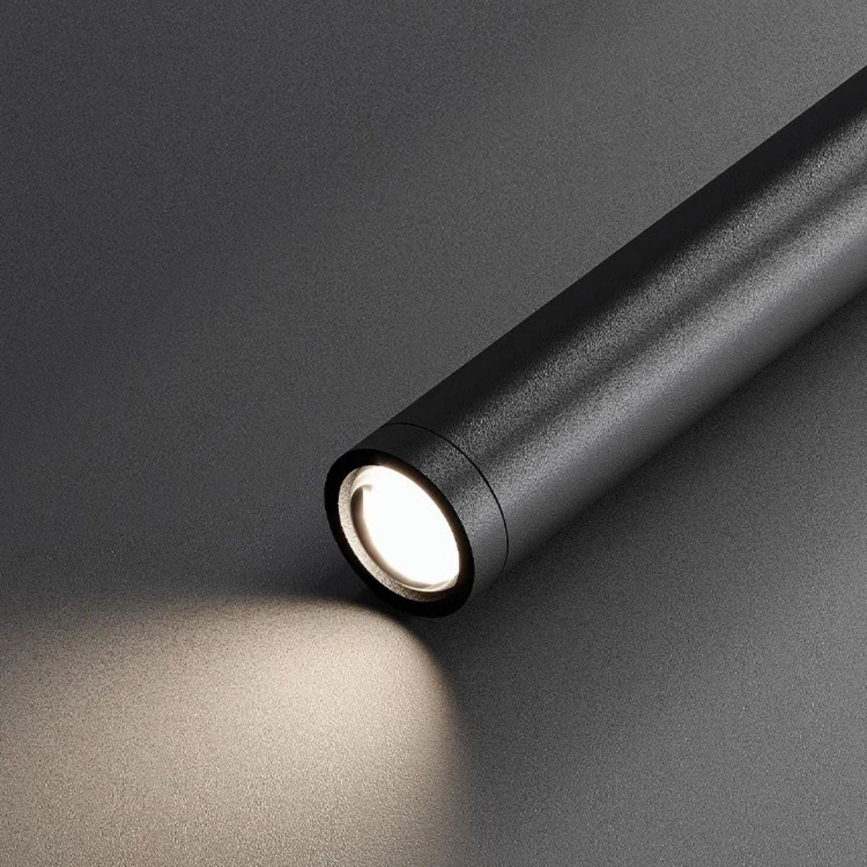 A sleek black cylindrical flashlight, resembling a cylindrical pendant light source, emits a soft beam of white light onto a smooth, dark surface, creating a gentle illuminated area in front of the device.