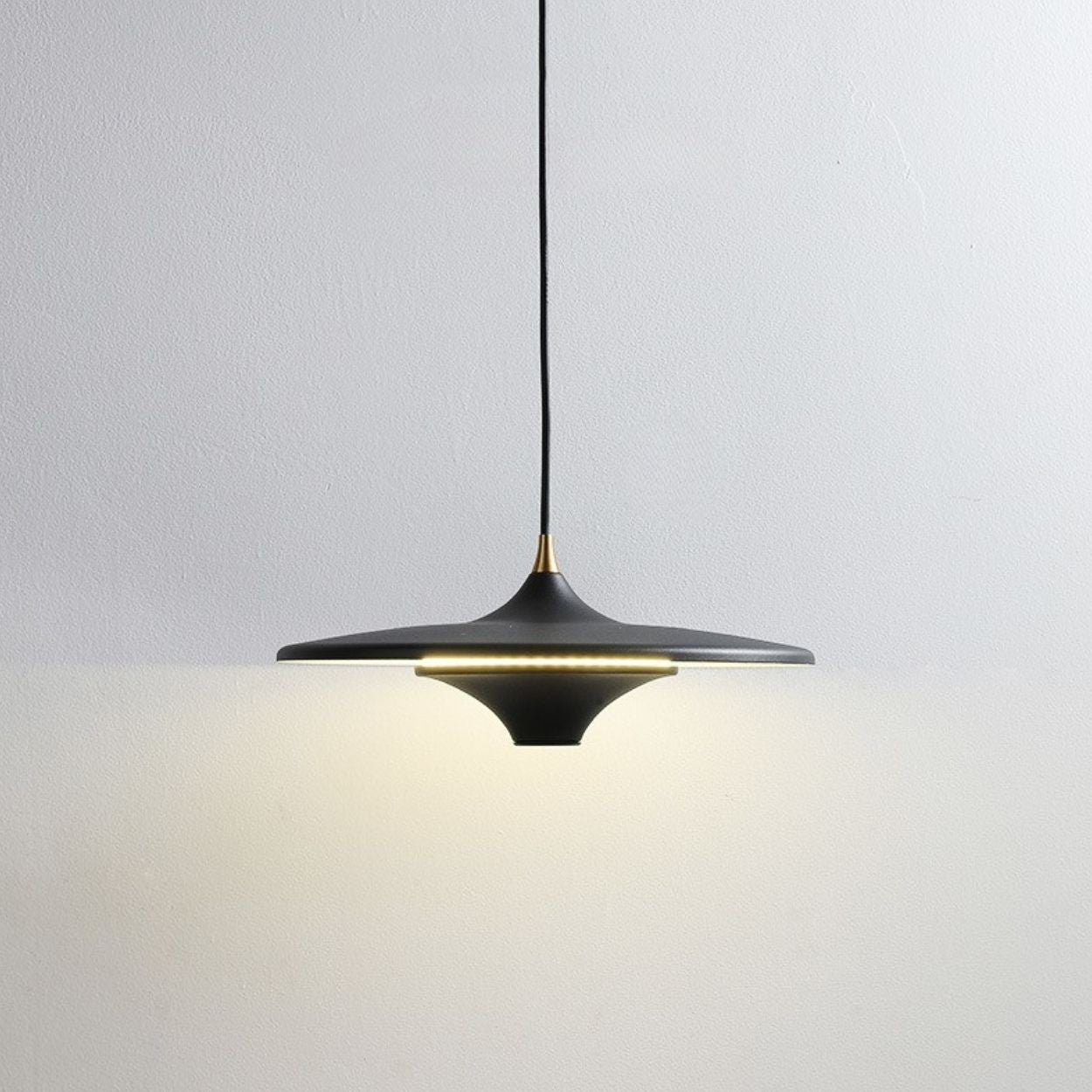 A modern black pendant light with a wide, flat shade hangs from the ceiling, casting a soft, warm glow on a plain white wall.